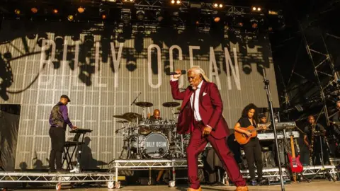 Camp Bestival Billy Ocean, a man with long white hair and a beard, is pictured on stage in a red suit. His band can be seen behind him. 
