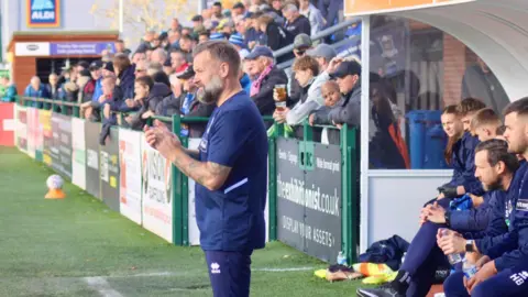 University of Chichester A man stands at the side of a football pitch. He is dressed in blue. He has a grey beard and is watching a game