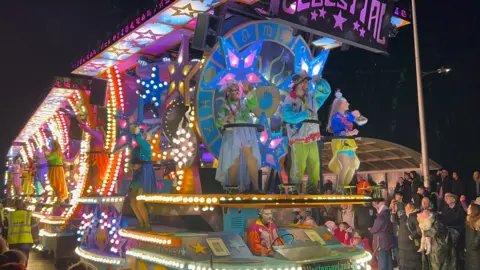 BBC A multicoloured illuminated carnival cart. There are three performers in costumes at the front of the cart and a line of others along the side. A crowd of people can be seen watching it.