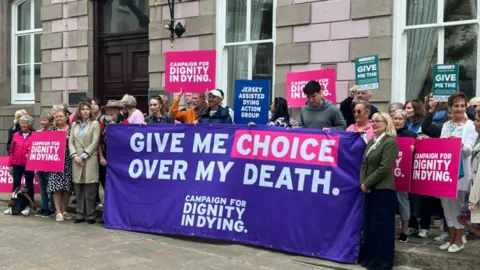 Campaigners for assisted dying holding a large purple "Give me choice over my death" and placards outside a grey building, the States Chamber.