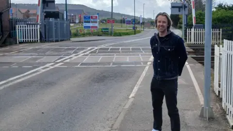 Tristan Learoyd is a man in his 30s wearing jeans. He is standing by a level crossing with a housing development behind him in the distance 