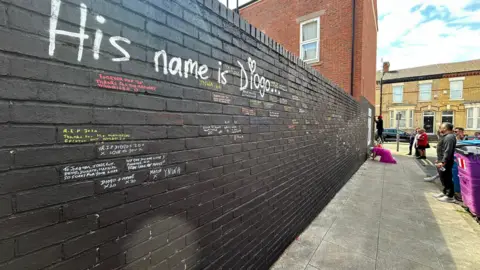 Tom Dunn/BBC A brick wall, painted black, with the words 'His name is Diogo' painted on in white lettering