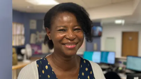 A woman with black hair smiles at the camera. She is wearing a cream-coloured cardigan over a dark blue dress patterned with what look like gold leaves. Behind her is a reception area with several computers.