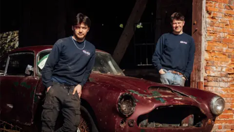 Two young men stand in front of a dilapidated red Aston Martin in a red-brick garage.