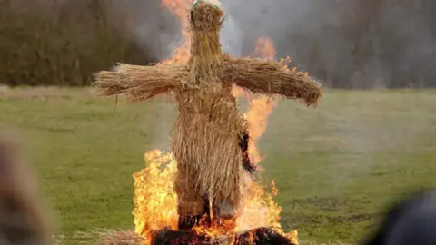 A figure made out of straw is set alight in a field. 