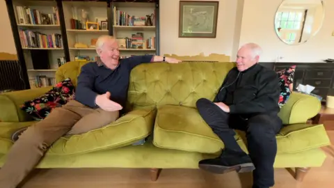 Nick Owen and Ian Phillipson sit on a couch and chat. Nick wears a black jacket and a black turtleneck sweater. Ian wears a blue sweater over a colorful shirt.