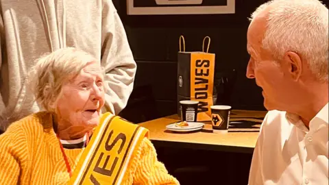 A woman with white hair wears an orange jumper, which is the same colour as the Wolves kit, with a sash that says Wolves. She is looking at and chatting to Steve Bull, who has white hair and is wearing a white shirt. Behind them is a table with a bag and some paper cups that have the Wolves logo on them. 