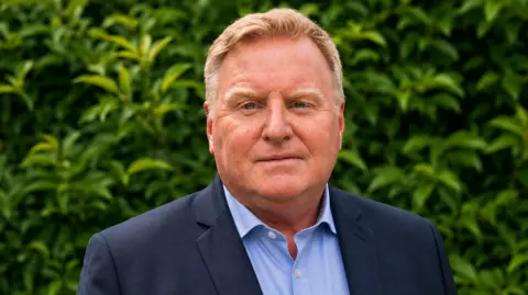 Andy Woolley has short blond hair. He wears a navy suit and a light blue shirt.