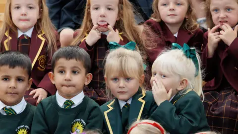 Four sets of twins - eight children in total - in school uniform, ahead of their first day at primary school. One child is sticking her tongue out at the camera, another is whispering to her sister. 