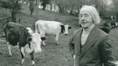 Fordhall A black and white picture of a man in a suit, standing in a field with cattle behind him
