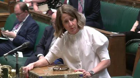 Liz Kendall speaking in Parliament. She is leaning on the despatch box, and in the process of answering a question. She is wearing a white blouse, and in front of her are her reading glasses.