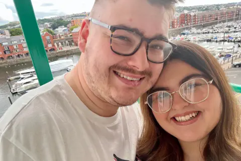 Ayla Jones A man in his 20s with a white t-shirt and dark tortoiseshell framed glasses leans his head onto Ayla Jones's. She has long brown hair and wears gold-framed glasses. It is a head and shoulders shot of them both smiling at the camera in a selfie pose. They appear to be high up with windows behind them and in the background the viewer can see Swansea Marina underneath with rows of converted warehouse flats and multiple small boats and yachts moored in the water.