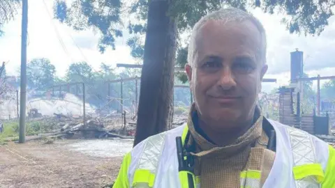 A man in a yellow firefighters jacket with bright yellow hi-vis vest over the top. He has closely cropped white hair and stands outside on a cloudy day with trees and fire damaged debris behind him.