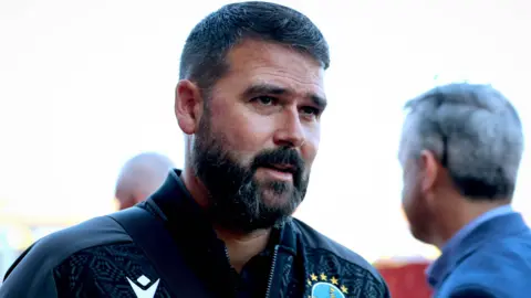 Inpho Linfield manager David Healy is looking to the right hand side of the frame. He Has dark brown hair and a dark brown beard. He is wearing a black zip up jersey.