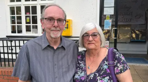 BBC / Grace McGrory The photo shows a man and a woman smiling at the camera. The man on the left has a grey striped shirt, and is wearing glasses. The woman on the right is wearing a floral top. They're standing in front of a white building. 