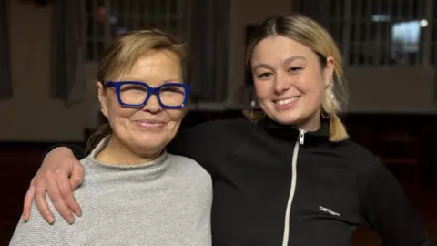 Eve and Levanna in a close-up shot smiling to camera with their arms around each other. Eve, the mother, is wearing a grey jumper and blue-framed glasses, and her daughter Levanna is in a black tracksuit top