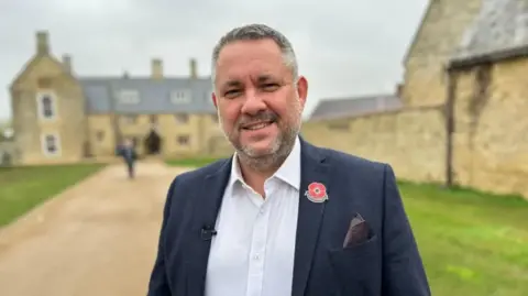 Kate Bradbrook/BBC A man with beard in a suit jacket and open necked while shirt smiling at the camera. He is standing in front of an old brick building