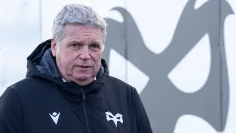 Lance Bradley wearing an Ospreys jacket in front of an Ospreys logo at Brewery Field