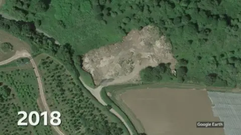 Google earth A satellite image with 2018 in white blocks on the bottom left of the image. The image shows the same scene as the previous one but there's now a large brown area on the right hand site where before there were fields with trees. The area appears to have been cleared and looks more like a builders yard as opposed to a countryside field.