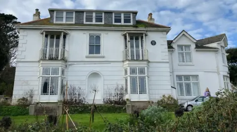 Talland House viewed from the front. It looks like a French-style villa with a more recent extension to the right. There are three storeys. There are bay doors on the ground floor and the windows on the first floor have balconies. The house is white. The garden extends in front of it.