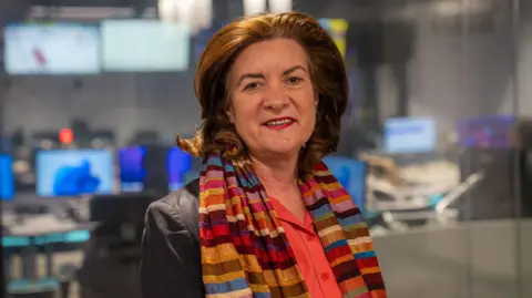 A photograph of Eluned Morgan at the BBC Wales Cardiff headquarters, with television screens in the blurred background. She is smiling and wearing a dark jacket, red shirt and multi-coloured scarf. 