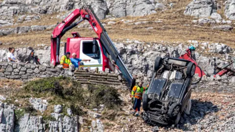 FPNW Upside down car pulled back to road above where a lorry with long arm and winch attempts to recover it