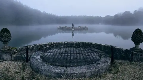 A mist-covered lake at Witley Park is viewed from a stone terrace. At the centre of the water is a platform topped by a statue of Neptune, marking the location of the underwater room hidden beneath the surface.