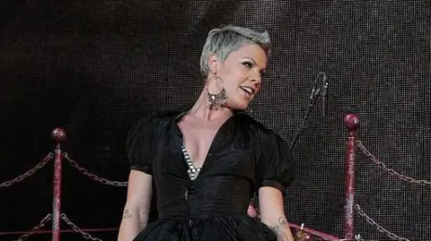 Getty Images Pink performs at Portman Road stadium in 2010. She is facing over her shoulder. She has large hooped earrings and wears a black jump suit.