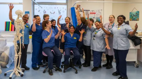Luton & Dunstable University Hospital A group of nurses, all wearing blue NHS outfits, with name labels on, in a hospital room. They have their arms up and are laughing and smiling. A skeleton is to the left, two are sitting on chairs and they are by a cubical. 