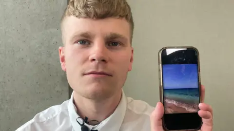 A young man with blonde hair and a stern expression, holding an iPhone which has the picture of a beach on it.