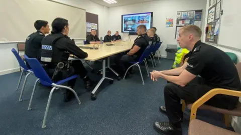 Thames Valley Police Officers being briefed as part of the operation in a meeting room at the police station. Some are sitting at a table, while others are sat on chairs away from it. There are various posters and leaflets on the walls and on cork boards. There is a computer monitor on and it appears to feature officers at other places on a live meeting.
