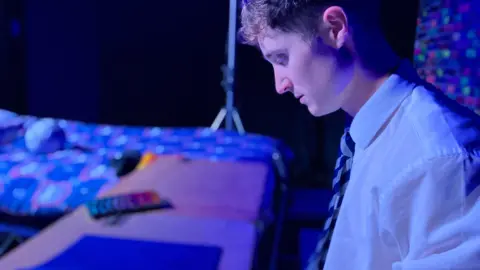 An actor on stage wearing a school shirt and tie sits at a desk, there is a bed in the background
