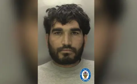 West Midlands Police A man with black hair and a black beard and moustache, wearing a grey t-shirt in front of a grey background