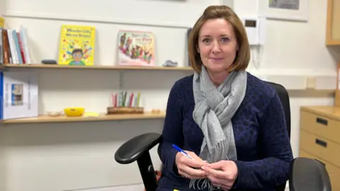 Vicki is smiling at the camera as she sits in a black chair by her desk. Behind are a few children's books on shelves. She has a dark blue with black dots jumper and a grey scarf. She's holding a blue biro pen in her hands and has short brown hair.