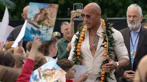 Joshua Falcon/BBC The Rock - a man wearing sunglasses and a cream-coloured top with flowers around his neck. He is smiling and looking down towards a group of schoolchildren.