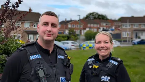 A male and a female police officer stand side-by-side in black uniform. They are stood on a housing estate and with properties and grass behind them. They are smiling at the camera. 
