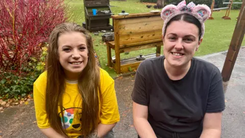 Two women pictured outdoors. The one on the left has long brown hair and is wearing a yellow Pudsey T-shirt. The one on the right has dark hair that's tied up and she is wearing a black T-shirt and pink Pudsey bear ears. Both of them are looking at the camera and smiling.