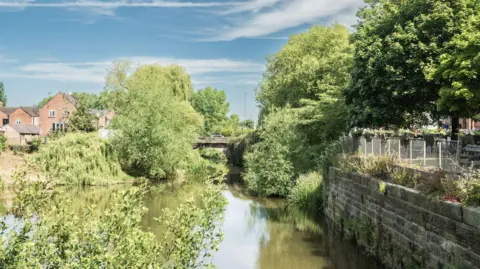 Getty Images A river seen from a boat on a sunny Day. A foot bridge can be seen ahead and a stone wall on the right bank, with trees either side and the backs of red brick houses to the left.