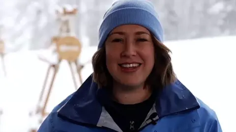BBC Lizzie Yarnold smiles at the camera during her interview with Radio Solent. She's wearing a blue jacket and beanie hat and behind her are the snowy slopes of Cortina.