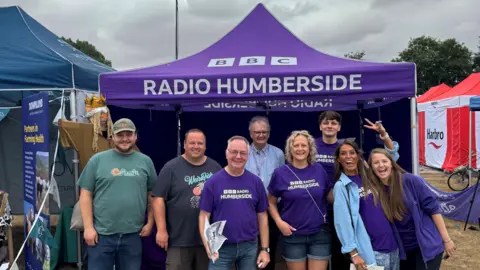 A group of five men and three woman stood in front of a purple BBC Radio Humberside tent. Some are wearing purple BBC Radio Humberside T-shirt. They are all smiling at the camera.