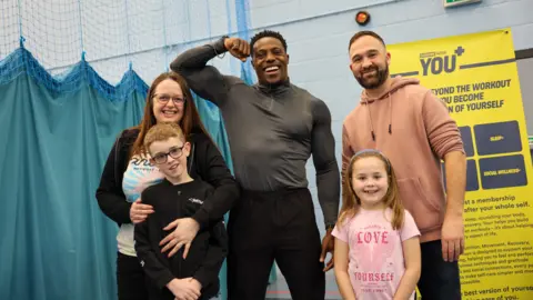 Everyone Active Harry Aikines-Aryeetey wearing black tracksuit bottoms and a grey long-sleeved gym shirt. He is posing with his bicep flexed and smiling at the camera. Beside him are a family with two small children, who are all smiling at the camera. They are standing in the Horfield Leisure Centre gym in front of a blue net curtain.