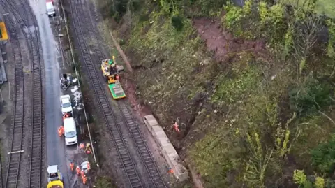 An overhead image of workers on a railway line who are repairing a damaged embankment on the right. Concrete blocks have been installed at the bottom of the embankment.