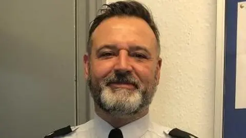 South Yorkshire Police Close-up image of Inspector Dodsworth. He is smiling slightly at the camera. He has brown short hair. He has a short beard which is a mix of brown, grey and white. He is wearing his police uniform.