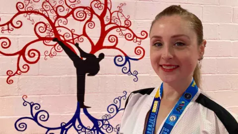 A woman wearing a white jacket with black stripes on the shoulders stands in front of a wall. She is wearing a blue lanyard. Behind her is the painting of a red and blue tree, featuring the silhouette of a man kicking his leg into the air.