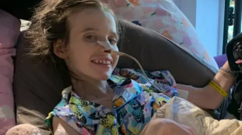 Family photo Hannah in her hospice bed, she is smiling at the camera, she has a medical tube attached to her nose