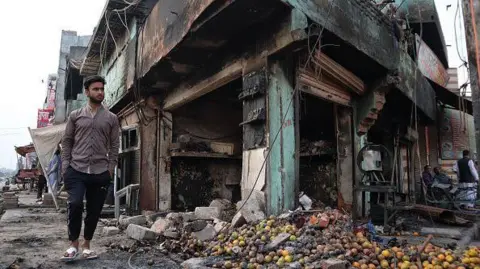 Getty Images A man walks besides a burnt fruit shop after a parking space was set on fire by a mob during riots in Chandbagh area of New Delhi, India on 29 February 2020.