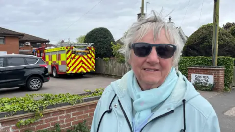 Andrew Turner/BBC Elaine Murphy is wearing sunglasses and has short hair. She has a light blue sky on and a matching light blue coat. She is standing in front of the centre's car park and a fire engine.