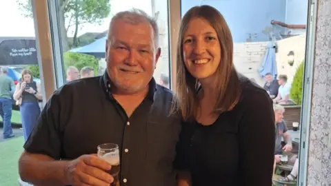 Neil Ainsworth A man on the left with grey hair and a dark shirt is holding a glass of drink in his hand. A woman with long brown hair and wearing a dark top is next to him.