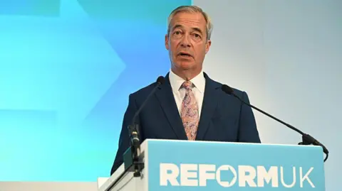 Nigel Farage standing a podium. He is wearing a navy suit and purple and beige tie.  He is standing in front of a light blue and white wall. The podium in front of him reads Reform UK, in the form of the party's logo.