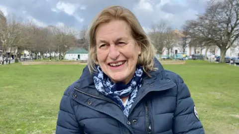 Assistant Health Minister Andy Howell - a woman in a scarf and blue parka jacket standing in front of a grass field 